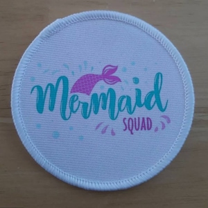 Pode incluir: Patch redondo de tecido branco com uma cauda de sereia rosa e o texto "Mermaid Squad" em letras azul turquesa.