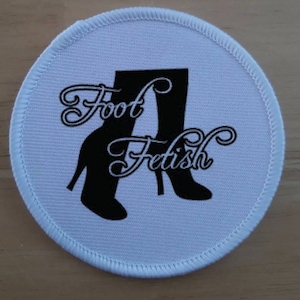 May include: White round patch with black embroidered text "Foot Fetish" and two black boot silhouettes.