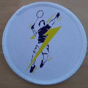 May include: White round patch with a yellow lightning bolt and a silhouette of a badminton player holding a racket. The player is wearing a dark blue shirt and shorts.