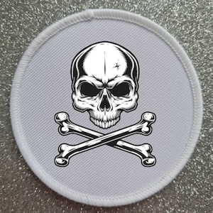 Pode incluir: Etiqueta redonda branca com um design de caveira e ossos cruzados em preto e branco. A caveira tem um design detalhado com uma rachadura na testa.