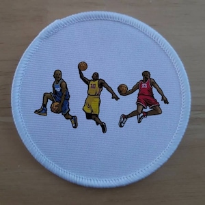 May include: A white round patch featuring three basketball players in action. The player on the left is wearing a blue jersey with the number 33, the player in the middle is wearing a yellow jersey with the number 10, and the player on the right is wearing a red jersey with the number 23.