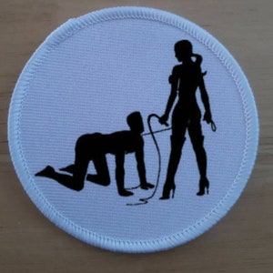 May include: A white round patch with a black silhouette of a person kneeling and another person standing with a whip. The person standing is holding the whip in their right hand and the person kneeling is looking up at the person standing.