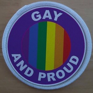 Könnte beinhalten: Ein runder lilafarbener Patch mit weißem Rand und dem Text "GAY AND PROUD" in Weiß. In der Mitte des Patches befindet sich ein regenbogenfarbener Kreis.