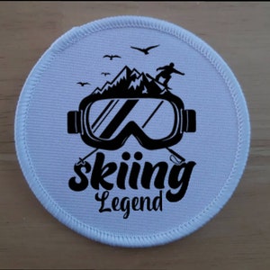 May include: White round patch with black ski goggles, mountains, and a skier. The text "skiing Legend" is printed below the goggles.