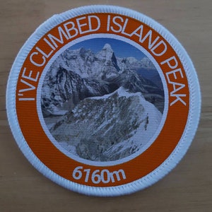 Könnte beinhalten: Ein runder weißer und orangefarbener Aufnäher mit dem Text "I've Climbed Island Peak 6160m" und einem Foto eines schneebedeckten Berges.