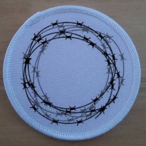 Può includere: Cerotto rotondo bianco con un design a filo spinato nero e grigio. Il filo spinato è disposto in un cerchio.