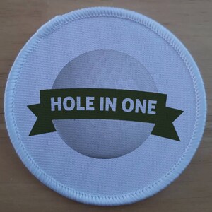 Puede incluir: Parche redondo de tela blanca con una cinta verde que dice "HOLE IN ONE" y una bola de golf blanca en el centro.