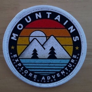 May include: White round patch with a black circle in the center. The circle has a retro sunset design with mountains, trees, and the text "MOUNTAINS EXPLORE ADVENTURE OUTDOOR" in a black circle.
