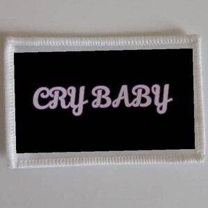 May include: White embroidered patch with a black background and the text "CRY BABY" in white with a purple outline.