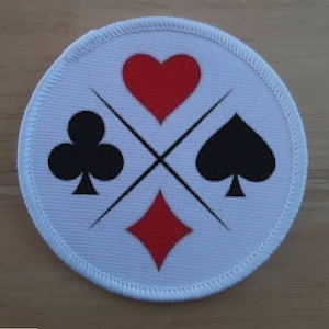 Pode incluir: Etiqueta redonda branca com quatro naipes de cartas de jogar pretos e vermelhos: um coração, uma espada, um trevo e um diamante. Os naipes estão dispostos em forma de cruz.