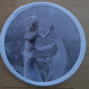 Pode incluir: Um remendo bordado branco com um design circular que apresenta uma mulher com um vestido branco e uma capa fluida. A mulher tem cabelo longo e fluente e levanta a mão no ar. O fundo é de cor azul claro.