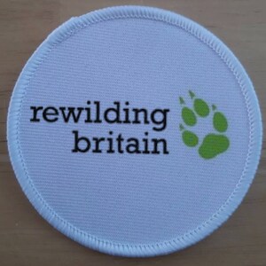 May include: White round fabric patch with black text that reads "rewilding britain" and a green paw print.