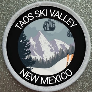 May include: A white circular patch with black text and a black circle in the centre. The text reads "Taos Ski Valley New Mexico". The black circle contains a graphic of a snowy mountain scene with ski lifts and skiers.
