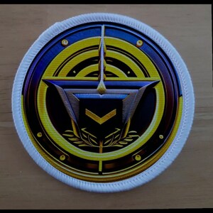 May include: A white circular patch with a gold, blue, and silver emblem. The emblem features a stylized star with a chevron inside, surrounded by a ring of gold and a smaller circle with a laurel wreath.