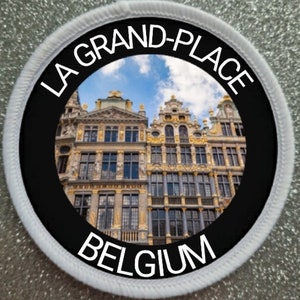 Puede incluir: Un parche redondo blanco y negro con el texto "LA GRAND-PLACE BÉLGICA" en letras blancas alrededor de una imagen circular de la Grand Place de Bruselas, Bélgica. La imagen muestra una vista de los edificios con un cielo azul y nubes blancas en el fondo.