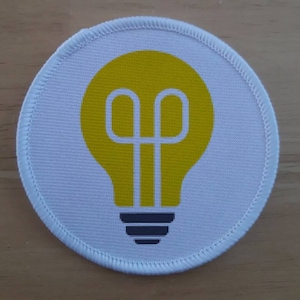 May include: A white round patch with a yellow light bulb design. The light bulb has a white outline and a black base.