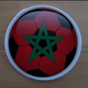 May include: A round, white fabric patch with a red and black football design. The football has a green five-pointed star in the centre.
