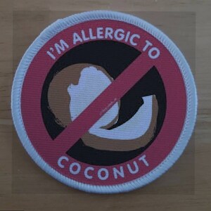 Pode incluir: Um remendo branco redondo com um círculo vermelho e uma linha vermelha diagonal que o atravessa. O círculo tem o texto "I'm allergic to coconut" em preto. Dentro do círculo há um coco marrom e branco.
