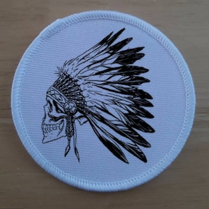 Op de afbeelding: Een ronde witte patch met een zwart-witte illustratie van een schedel die een hoofdtooi met veren draagt.