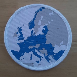 Puede incluir: Un parche circular blanco con un mapa de Europa azul y gris impreso en él. El mapa muestra las fronteras de todos los países de Europa. El parche tiene un borde blanco alrededor del borde.