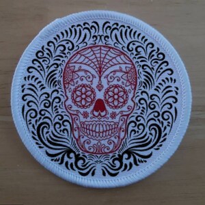 以下が含まれることがあります： 赤色のシュガースカルデザインが施された、丸い白いパッチ。スカルは花柄とウェブパターンで精巧に装飾されています。黒い渦巻き模様の装飾要素がスカルを囲み、死者の日のテーマを作り出しています。