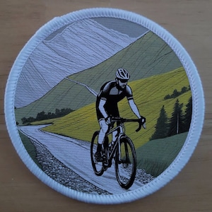 Op de afbeelding: Een witte, ronde patch met een zwart-witte illustratie van een fietser die een fiets rijdt op een kronkelende weg door een groen en geel landschap. De fietser draagt een zwart shirt en helm. De achtergrond toont een bergketen met een witte, bewolkte lucht.