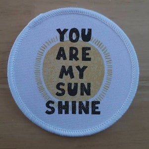 Peut inclure: Patch rond en tissu blanc avec un graphique de soleil jaune et le texte "YOU ARE MY SUNSHINE" en noir.