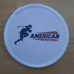 Könnte beinhalten: Ein runder, weißer Aufnäher mit einer gestickten Umrandung. Er zeigt einen Footballspieler in Bewegung als Silhouette, mit dem Schriftzug "AMERICAN FOOTBALL" in Rot und Schwarz.