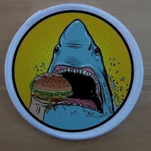 May include: A white, round patch featuring a blue shark taking a bite out of a hamburger. The shark has a white mouth with sharp teeth. The hamburger is yellow, brown, and green. The background is yellow with a comic book style pattern.