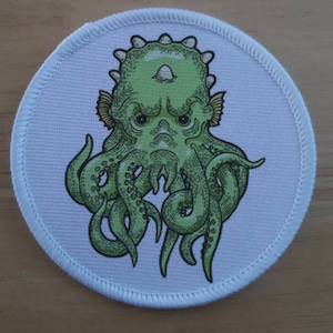Op de afbeelding: Een witte ronde patch met een groene cartoon octopus met een chagrijnige uitdrukking. De octopus heeft een stekelig hoofd en grote oren.