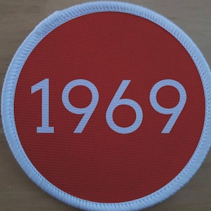 Könnte beinhalten: Ein roter runder Patch mit weißem Rand und der Zahl 1969 in weiß gedruckt.