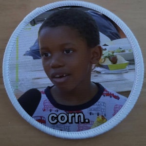 May include: A white round patch with a white border. The patch features a photo of a young boy wearing a red and white patterned shirt. The text "corn." is printed in black letters at the bottom of the patch.