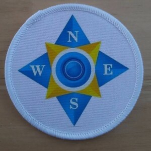以下が含まれることがあります： 青と黄色のコンパスデザインが施された白い布のパッチ。コンパスは北、南、東、西を指しています。