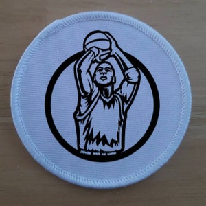 May include: A circular, white patch featuring a black illustration of a basketball player in the act of shooting. The player is enclosed within a black circle, with a white stitched edge.