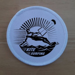May include: A white round patch with a black and white illustration of a person kite surfing. The text "KITE SURFING" is printed below the illustration.