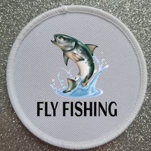 Op de afbeelding: Ronde witte patch met een kleurrijke illustratie van een vis die uit het water springt. De tekst "FLY FISHING" is in zwart onder de vis gedrukt.