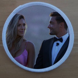 May include: A white circular patch with a photo of a couple looking at each other. The woman is wearing a pink dress and the man is wearing a black suit with a black bow tie.