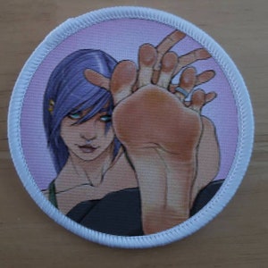 May include: A white round patch with a purple-haired person with a ring on their finger, holding their foot up in the air. The background is a light pink.