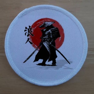 May include: White round patch with a black and white image of a samurai warrior standing in front of a red sun. The kanji character "忍" is visible on the left side of the image.