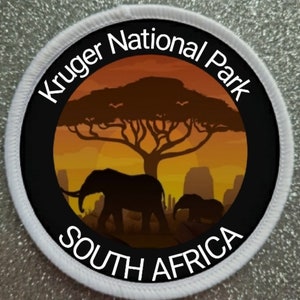 Könnte beinhalten: Ein weißer runder Patch mit schwarzem Text, der "Kruger National Park South Africa" lautet. Der Patch zeigt eine Silhouette eines Akazienbaums mit zwei Elefanten, die davor laufen. Der Hintergrund ist ein Sonnenuntergang in orange und gelben Farbtönen.