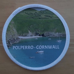 May include: A white round patch with a photo of a cove with a sandy beach and blue water. The text "POLPERRO - CORNWALL" is printed in white on the patch.