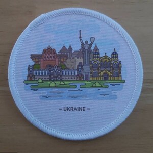 May include: A white round patch with a colorful illustration of a cityscape with buildings and a statue. The text "UKRAINE" is printed in black at the bottom of the patch.