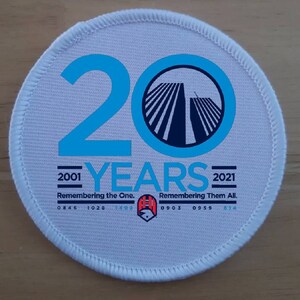 以下が含まれることがあります： 青い数字の20と、青と黒の双子塔のシルエットが描かれた白い丸いパッチ。塔の下には「2001 YEARS 2021」という文字が印刷されています。「Remembering the One.」と「Remembering Them All.」という文字が年の下に印刷されています。「0846 1028 1402 0903 0959 614」という文字がテキストの下に印刷されています。