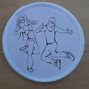 May include: A white round patch with a black line drawing of two dancers. The female dancer is on the left and the male dancer is on the right. The female dancer is wearing a tank top and pants. The male dancer is wearing a tank top, pants, and a hat.