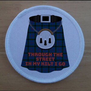 Puede incluir: Un parche redondo blanco con una falda escocesa azul, verde y roja a cuadros, un sporran blanco y el texto "Through the street in my kilt I go" en naranja.