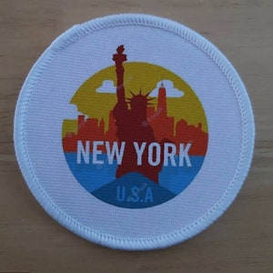 May include: A white circular fabric patch featuring a colourful image of the Statue of Liberty and the New York City skyline. The text "NEW YORK" and "U.S.A." are printed on the patch.
