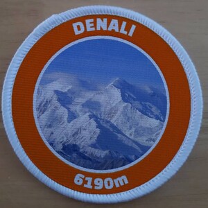 May include: A white and orange round patch with a picture of a snow-capped mountain range. The text "DENALI" is at the top of the patch and "6190m" is at the bottom.