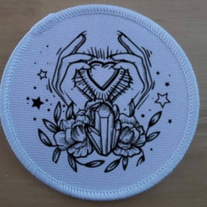 Op de afbeelding: Een ronde witte patch met een zwart-witte illustratie van twee handen die een hartvormig kristal vasthouden. Het kristal wordt omringd door bloemen en sterren.