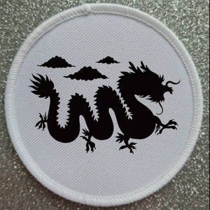 May include: A white round fabric patch with a black silhouette of a dragon with clouds in the background.