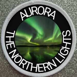 Può includere: Una toppa circolare bianca con testo nero che recita "Aurora The Northern Lights" attorno a una foto dell'aurora boreale verde riflessa in uno specchio d'acqua.
